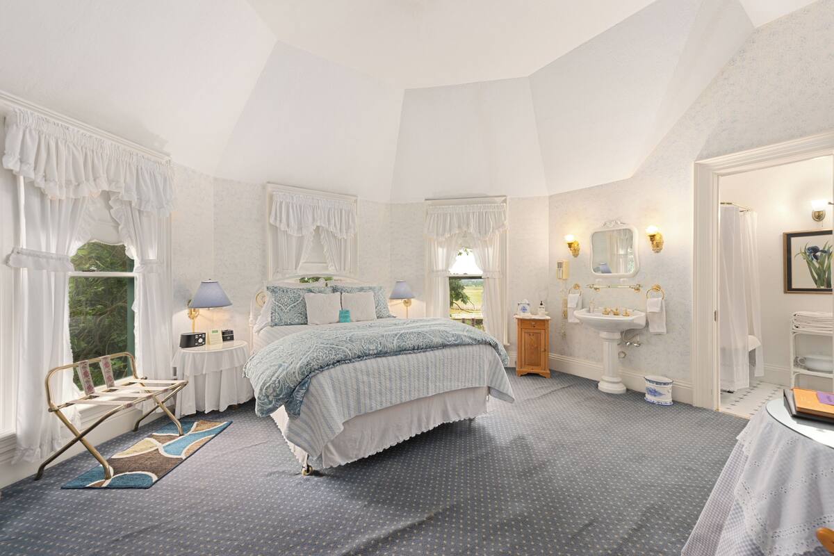 A bedroom with a large bed, a bedside table with a lamp, a nightstand, a window with curtains, a bathroom with a sink and mirror, and a view of the outdoors.