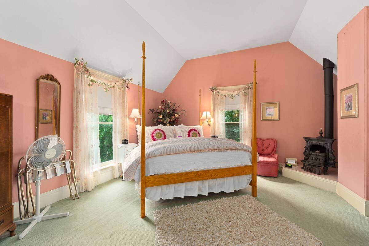 A bedroom with a four-poster bed, a standing fan, a fireplace, and a pink wall.