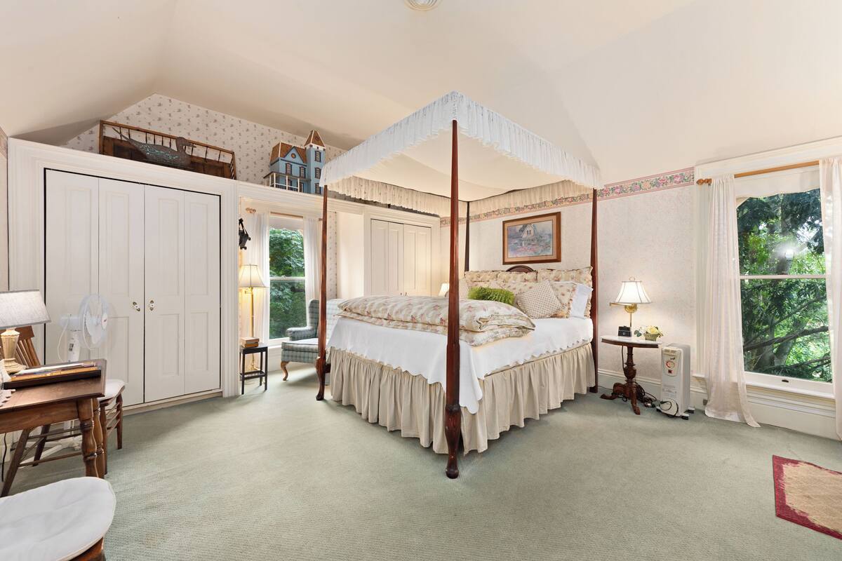 A four-poster canopy bed with a floral bedspread, a desk with a lamp, and a view of a blue house through a window.