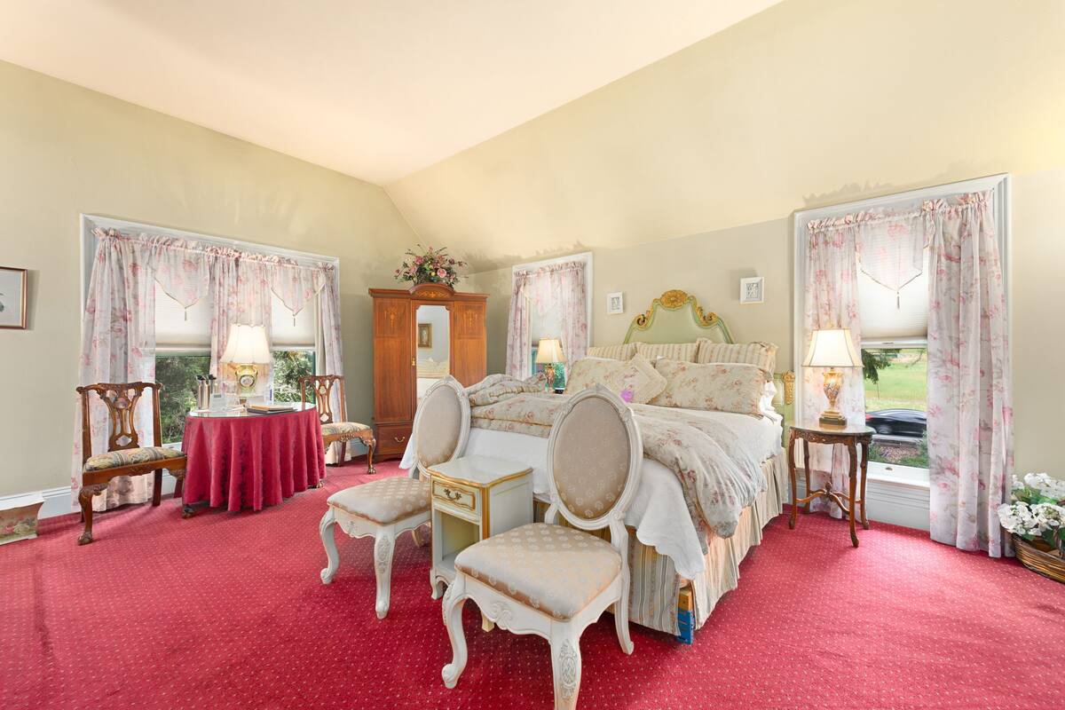 A bedroom with a large bed, two bedside tables, a chair, a small table, and a window with curtains.