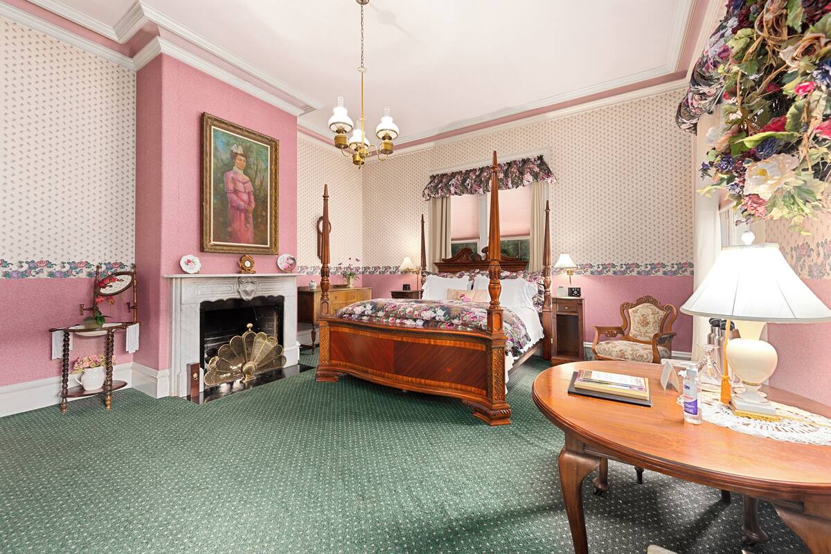 A classic bedroom with a four-poster bed, a fireplace, a dining table, and a chandelier.