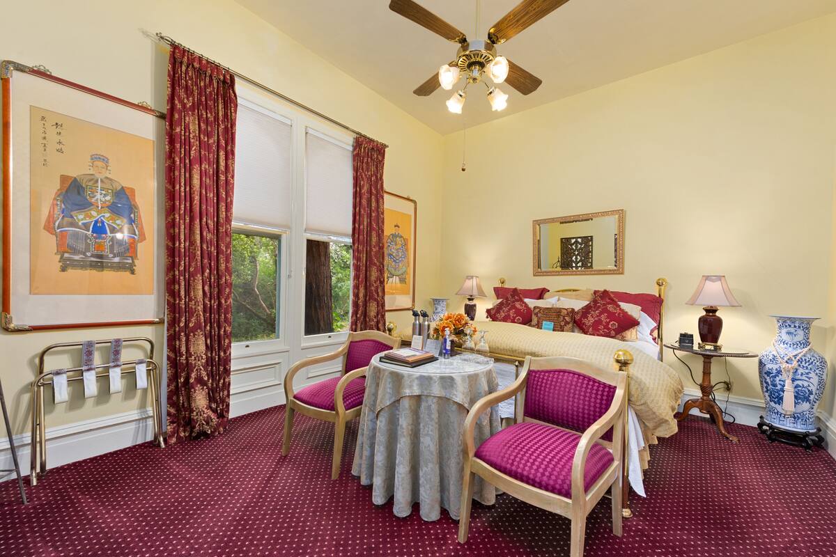 A traditional room with a bed, a round table with chairs, a ceiling fan, and a large vase.