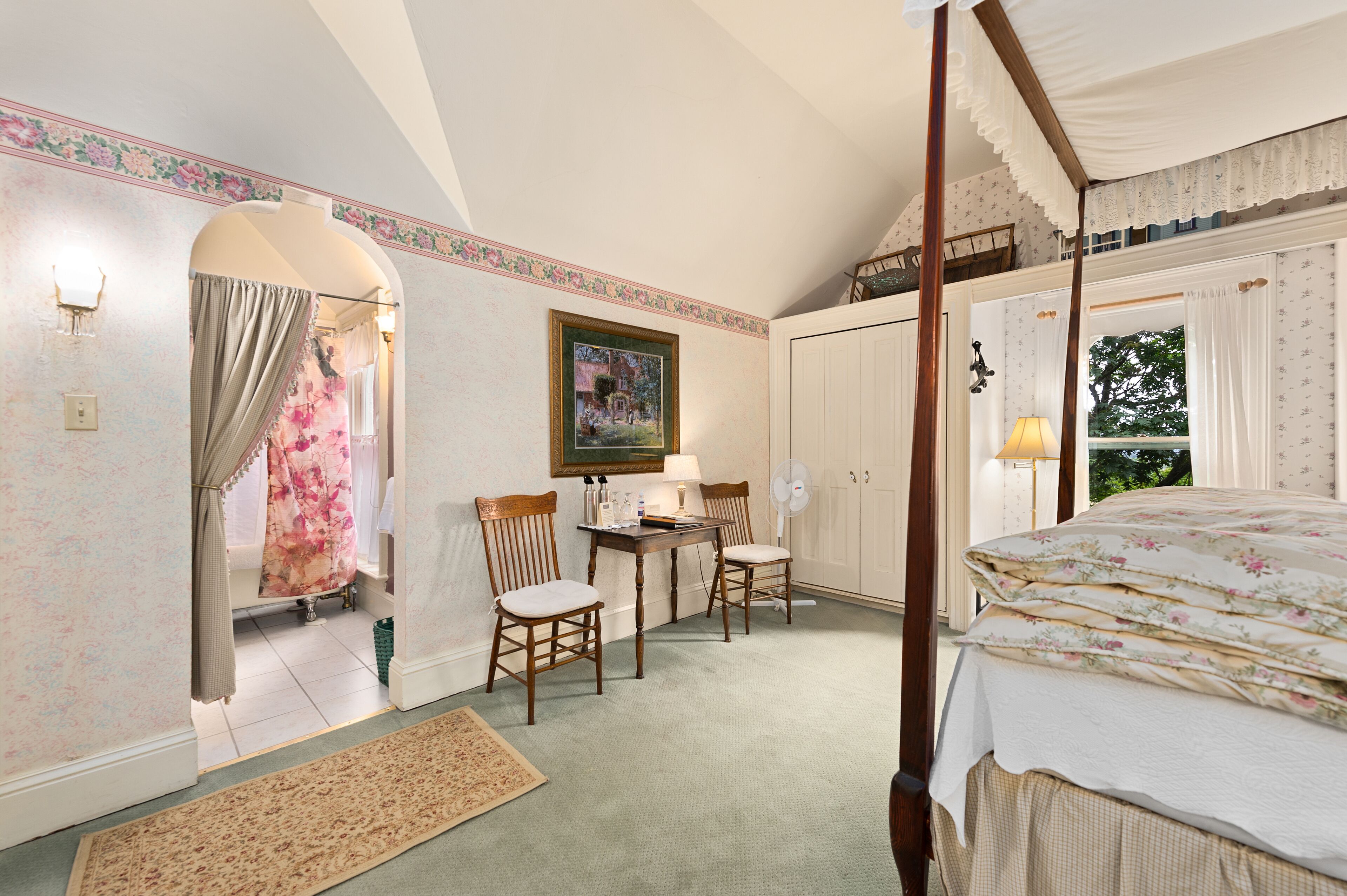 A bedroom with a four-poster bed, a desk with chairs, a painting on the wall, and a view of the outdoors through a window.