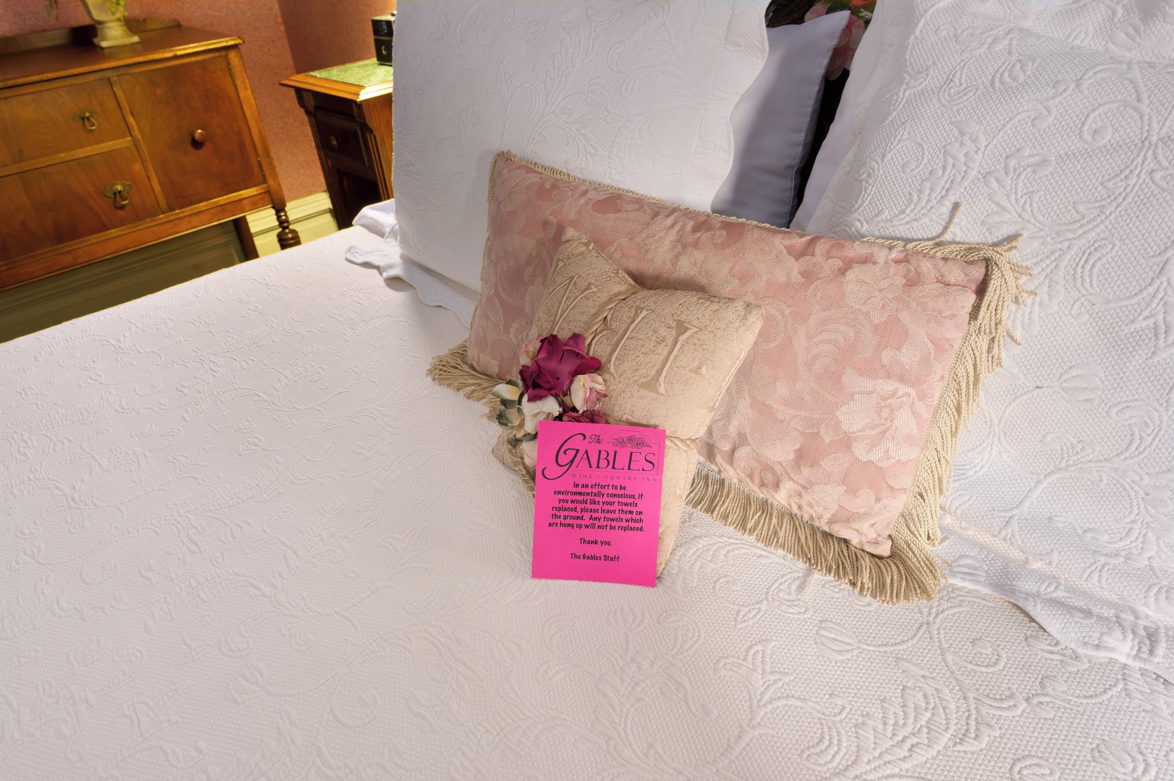 Un lit bien fait, avec un oreiller rose et beige, une étiquette indiquant « The Gables », et une table de chevet en bois dotée de tiroirs.