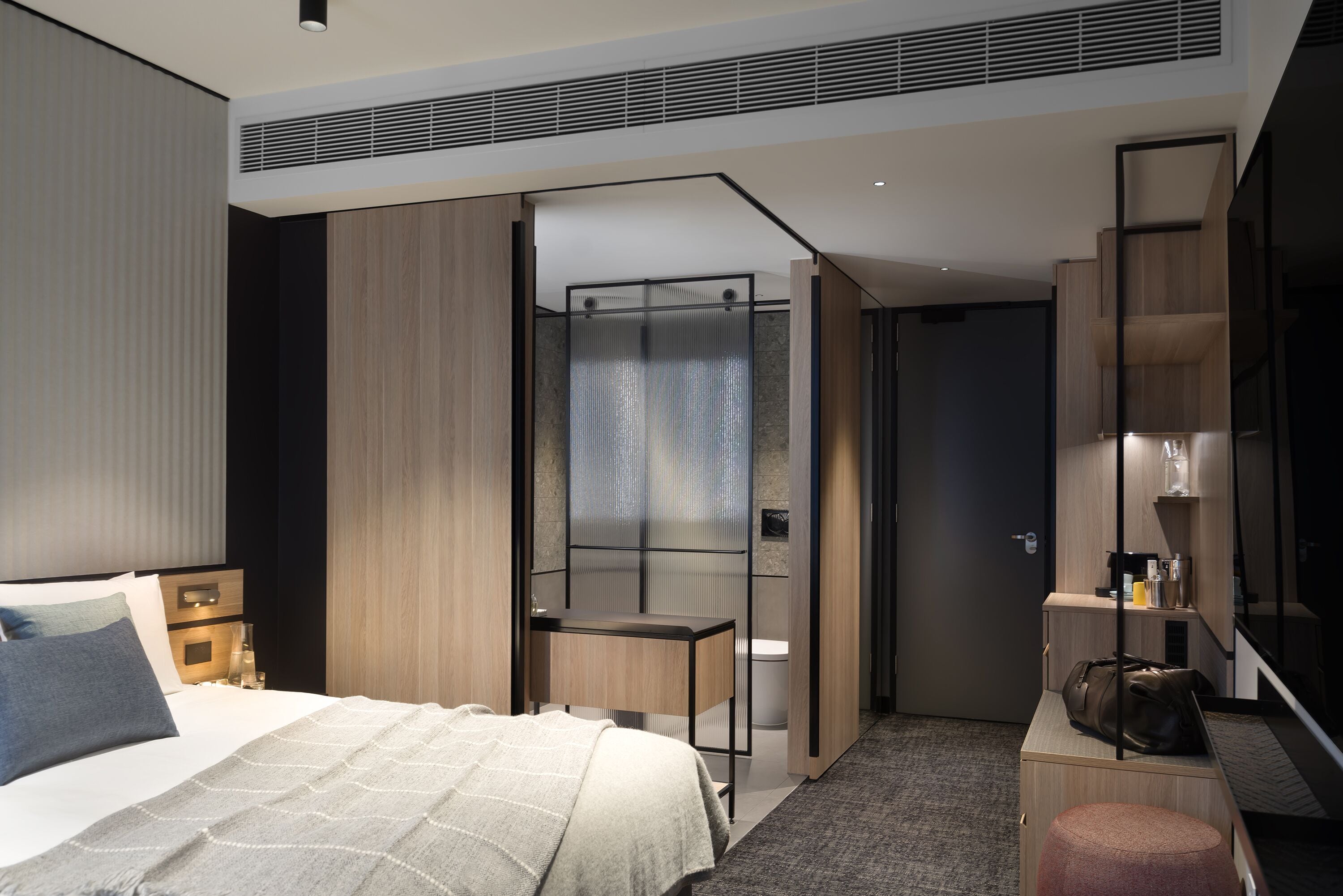 A modern hotel room with a bed, bedside table, and a desk with a chair. There is a bathroom visible through a glass partition.