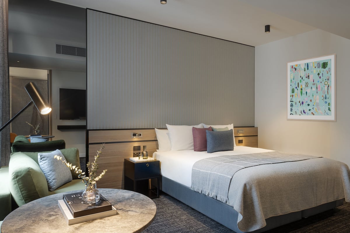 Habitación de hotel moderna con una cama bien hecha, una mesita de noche con lámpara, una mesita redonda con un libro y un jarrón, y un cuadro abstracto grande en la pared.