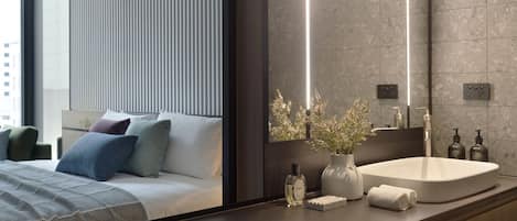 A modern hotel room with a large bed, a bedside table with a vase of flowers, a white sink, and a large mirror.
