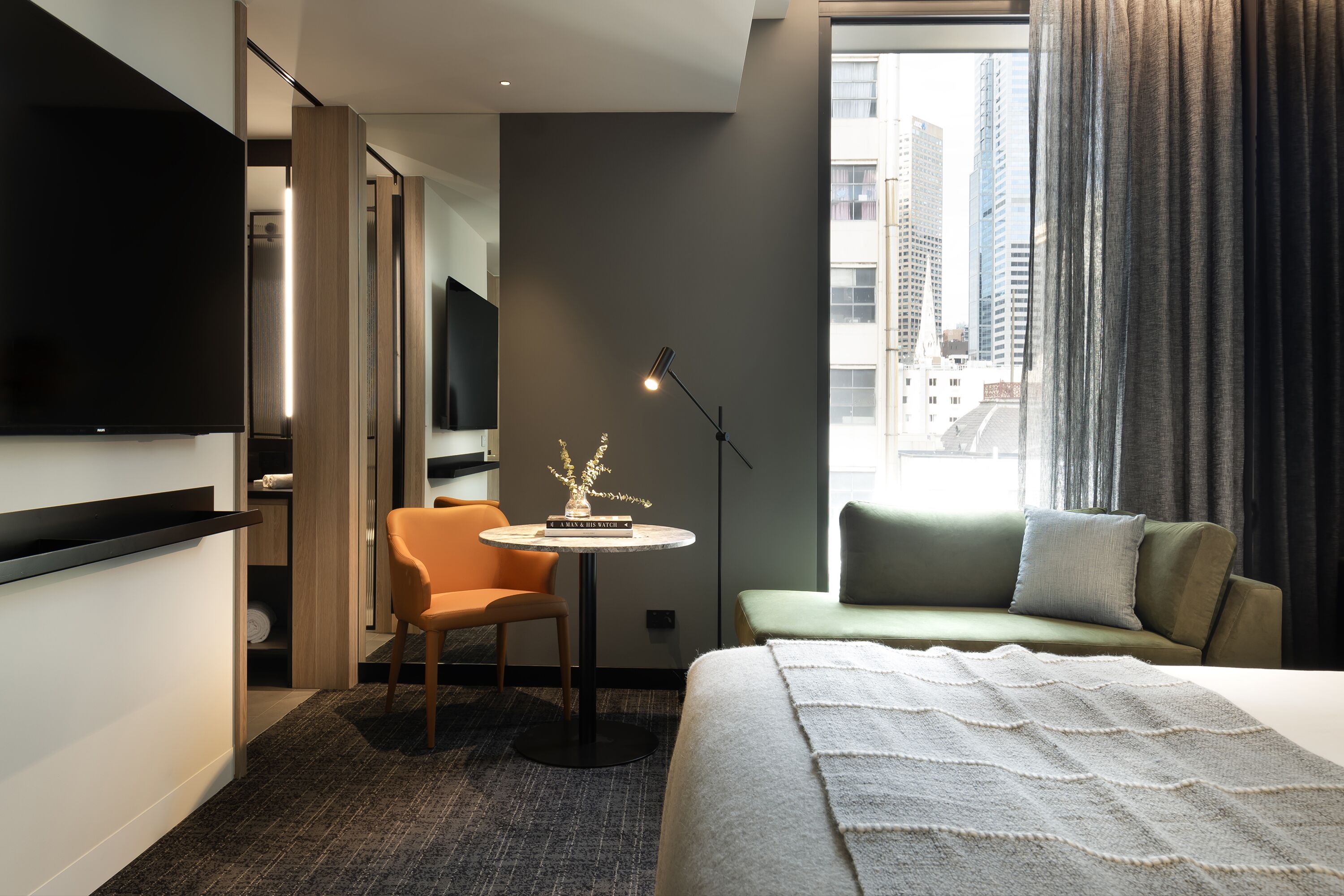 A modern hotel room with a bed, a seating area, a TV, a small table with a decorative item, and a view of the city through the window.
