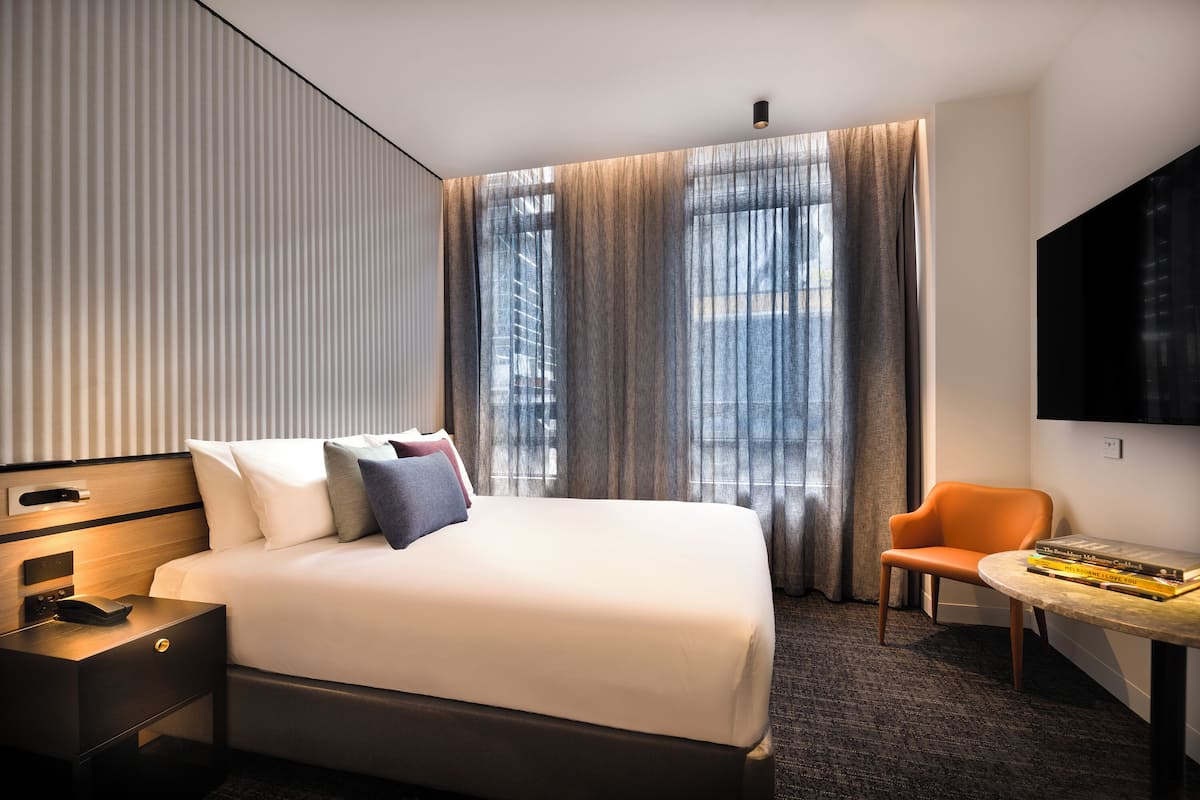 Una habitación de hotel moderna con una cama grande, una mesita de noche con teléfono, una silla, una pequeña mesa con libros y un televisor en la pared.