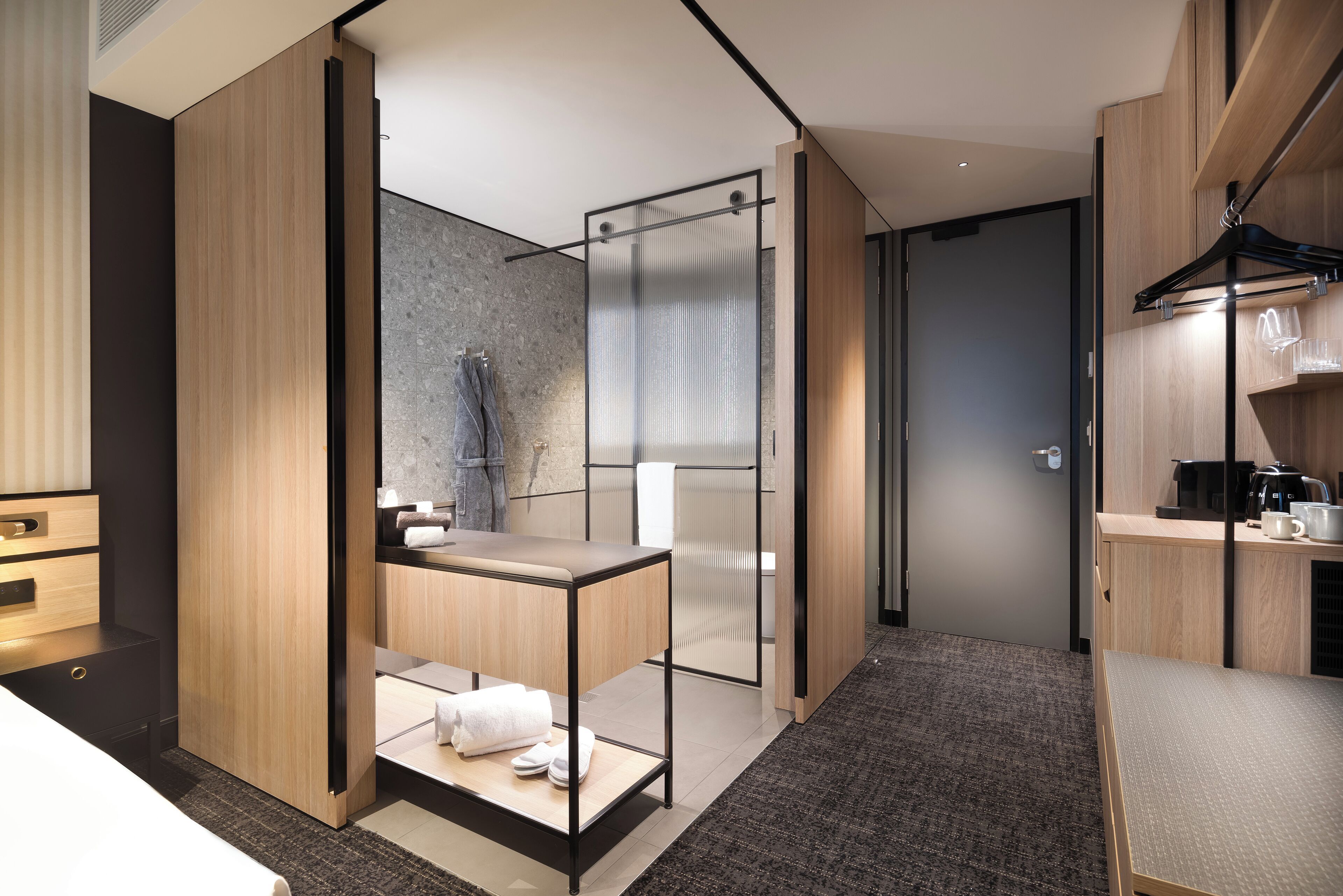 A modern hotel room with a wooden wardrobe, a glass shower enclosure, a small table with toiletries, and a coffee machine on the right side.