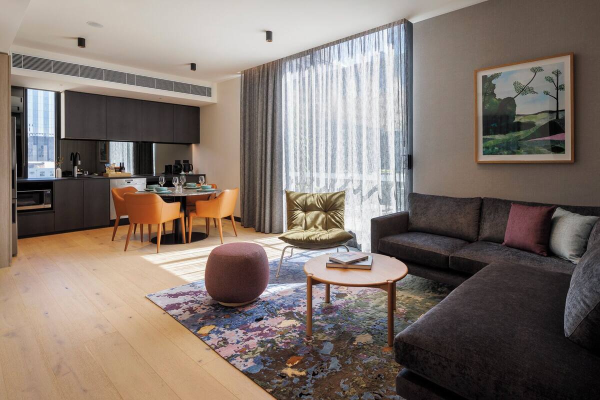 Una sala de estar moderna con un sofá, una mesa de centro, una zona de comedor con sillas, una cocina al fondo y un amplio ventanal con cortinas.