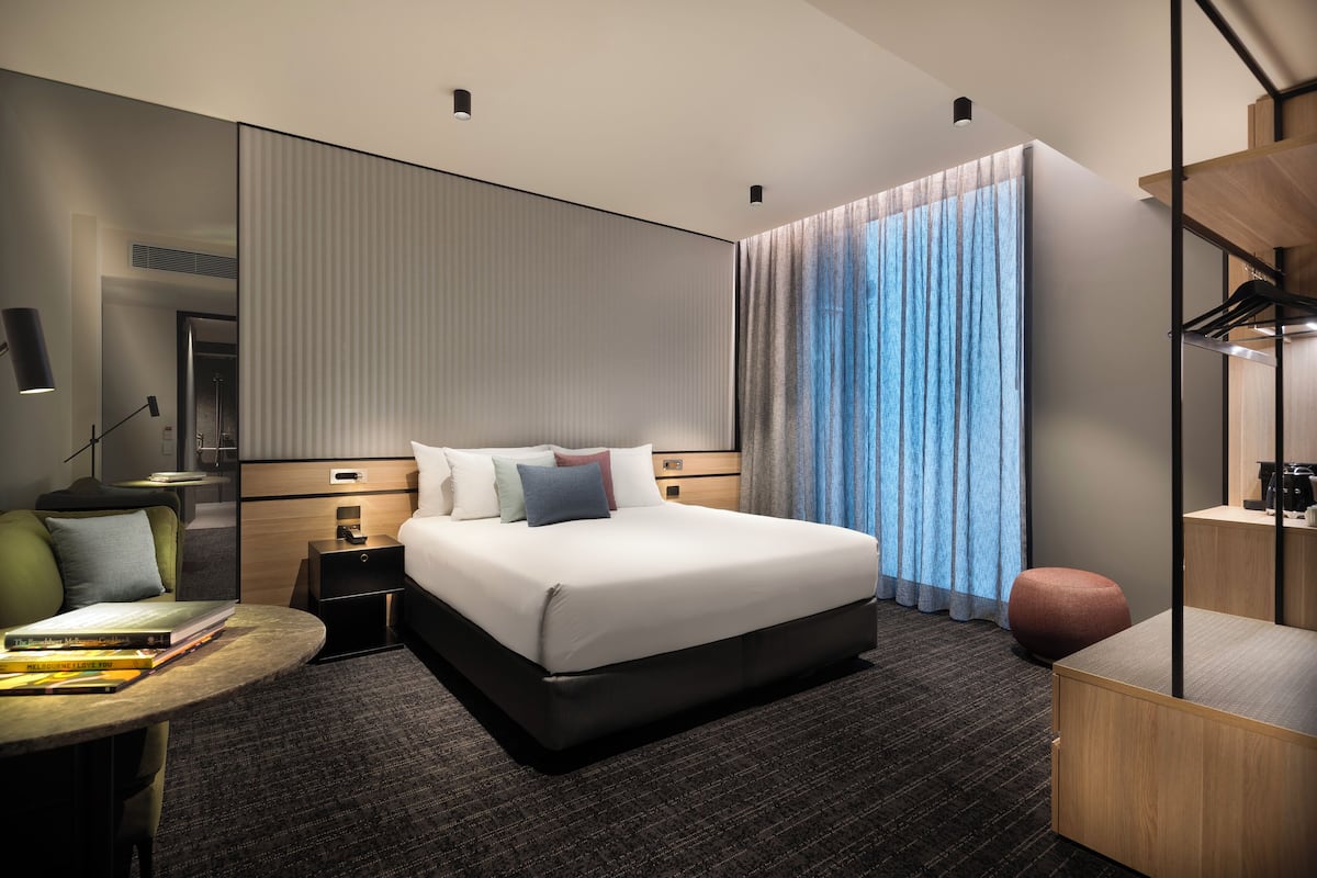 Una habitación de hotel moderna con una cama grande, una mesita de noche, un escritorio y un amplio ventanal con cortinas.