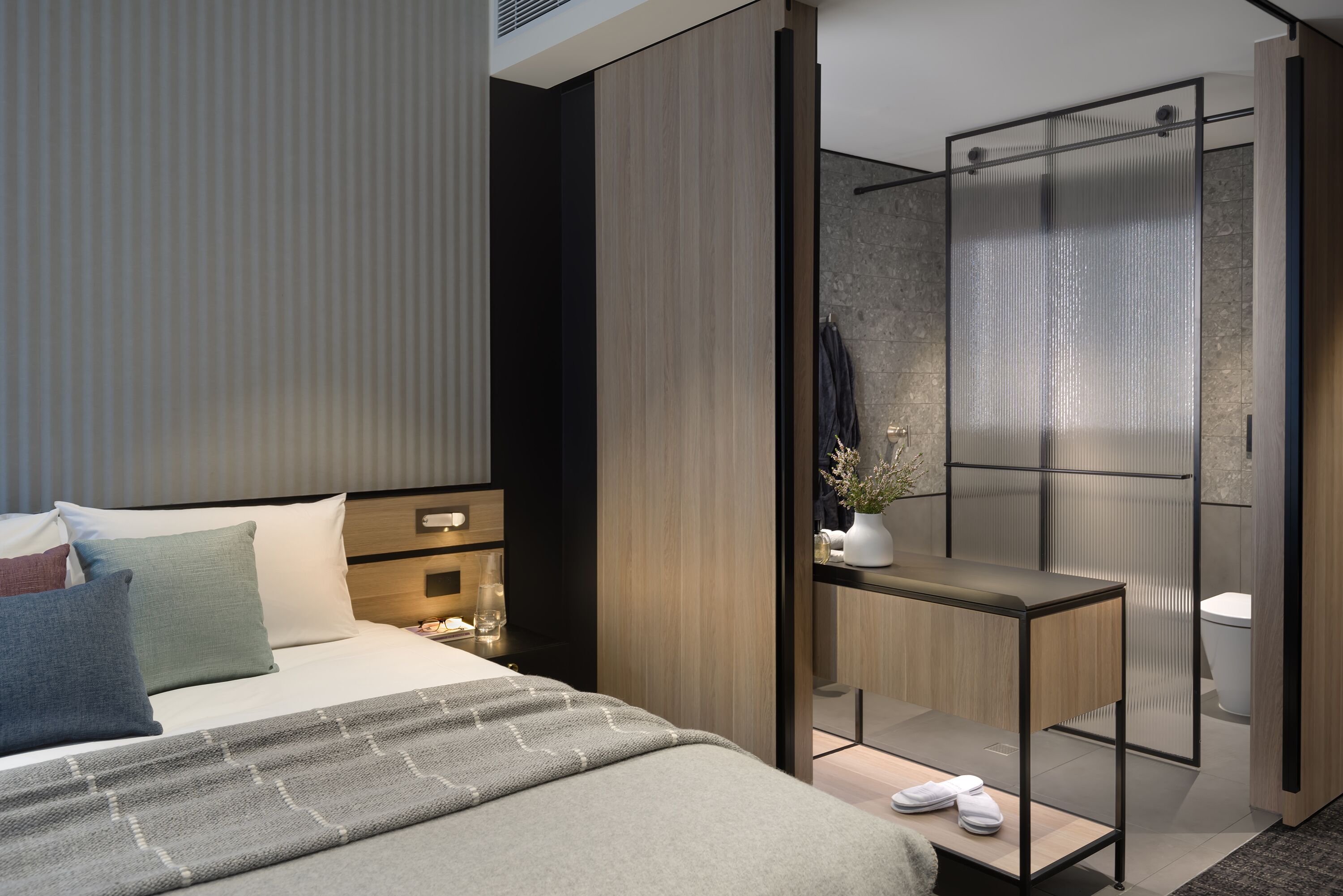 A modern hotel room with a bed, bedside table, and a shower area with a glass door.