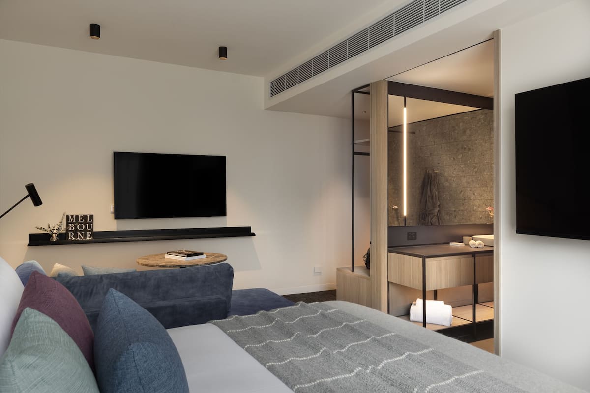 Un dormitorio moderno con una cama grande, un televisor en la pared, una mesita de noche con lámpara y vistas al baño.