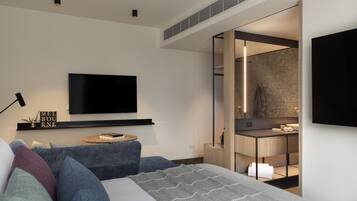 A modern bedroom with a large bed, a TV mounted on the wall, a bedside table with a lamp, and a view into the bathroom.