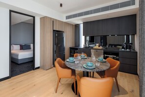 A modern kitchen with dark cabinetry, a dining table with orange chairs, and a separate bedroom with a bed and a nightstand. - The Howey (Melbourne)