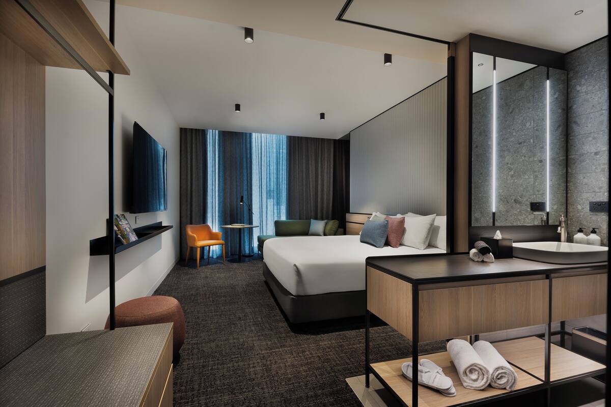 Una habitación de hotel moderna con una cama grande, un escritorio, una silla, un televisor, un baño con espejo y vistas a la ciudad.