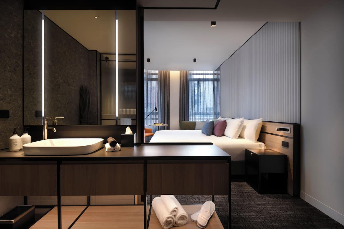 Una habitación de hotel moderna con una cama grande, una mesita de noche, un tocador, un espejo amplio, un baño con lavabo y una ventana con cortinas.