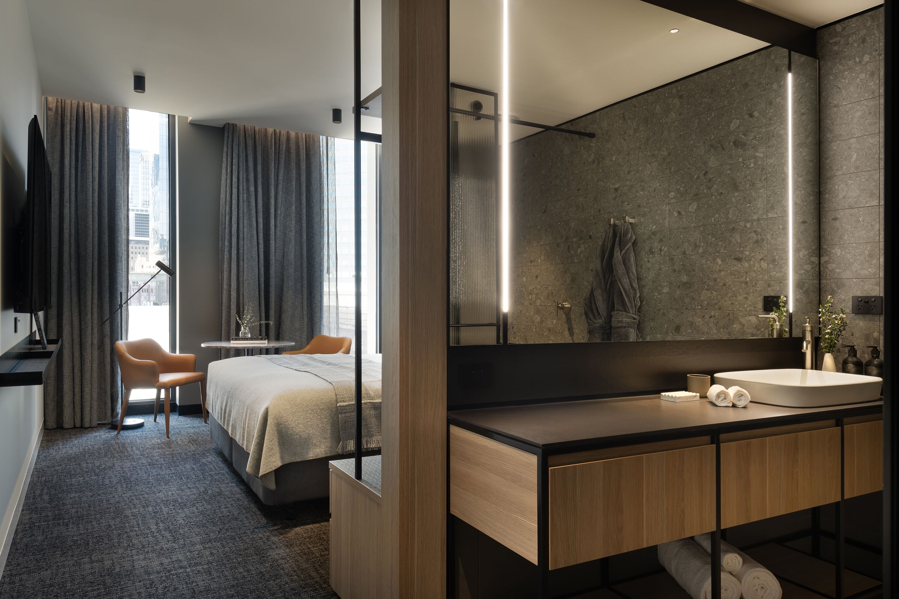 A modern hotel room with a large bed, a sitting area with a chair, a bathroom with a large mirror, and a view of the city through the window.