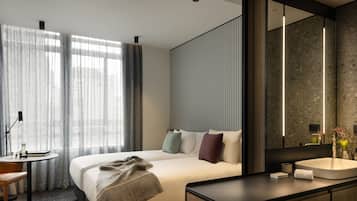 Una habitación de hotel moderna con una cama grande, una mesita de noche con lámpara, un escritorio pequeño y un baño con un espejo grande y lavamanos.