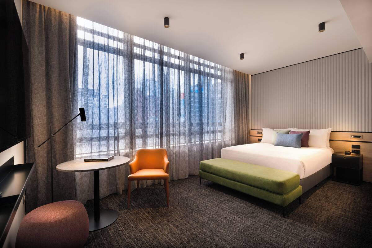 Habitación de hotel moderna con una cama grande, un reposapiés verde, una mesita redonda pequeña, una silla y un escritorio con lámpara.