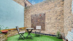 Un patio con muro de ladrillo, dos sillas, una mesa pequeña y un muro decorativo con adornos colgantes.