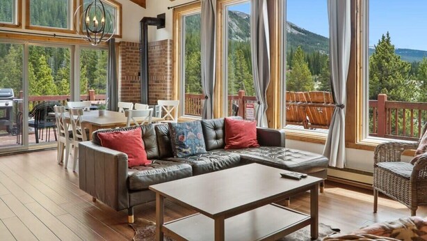 Ein gemütliches Wohnzimmer mit einer Eckcouch, einem Holztisch und einem Essbereich mit Tisch und Stühlen. Große Fenster bieten Blick auf Berge und Wald.