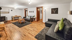 Ein gemütliches Wohnzimmer mit Holzboden, einer schwarzen Ledersofa, einem Couchtisch aus Holz und einem grauen Teppich. Es gibt einen Essbereich mit Holztisch und Stühlen sowie eine kleine Küchenzeile mit Herd und Schränken.