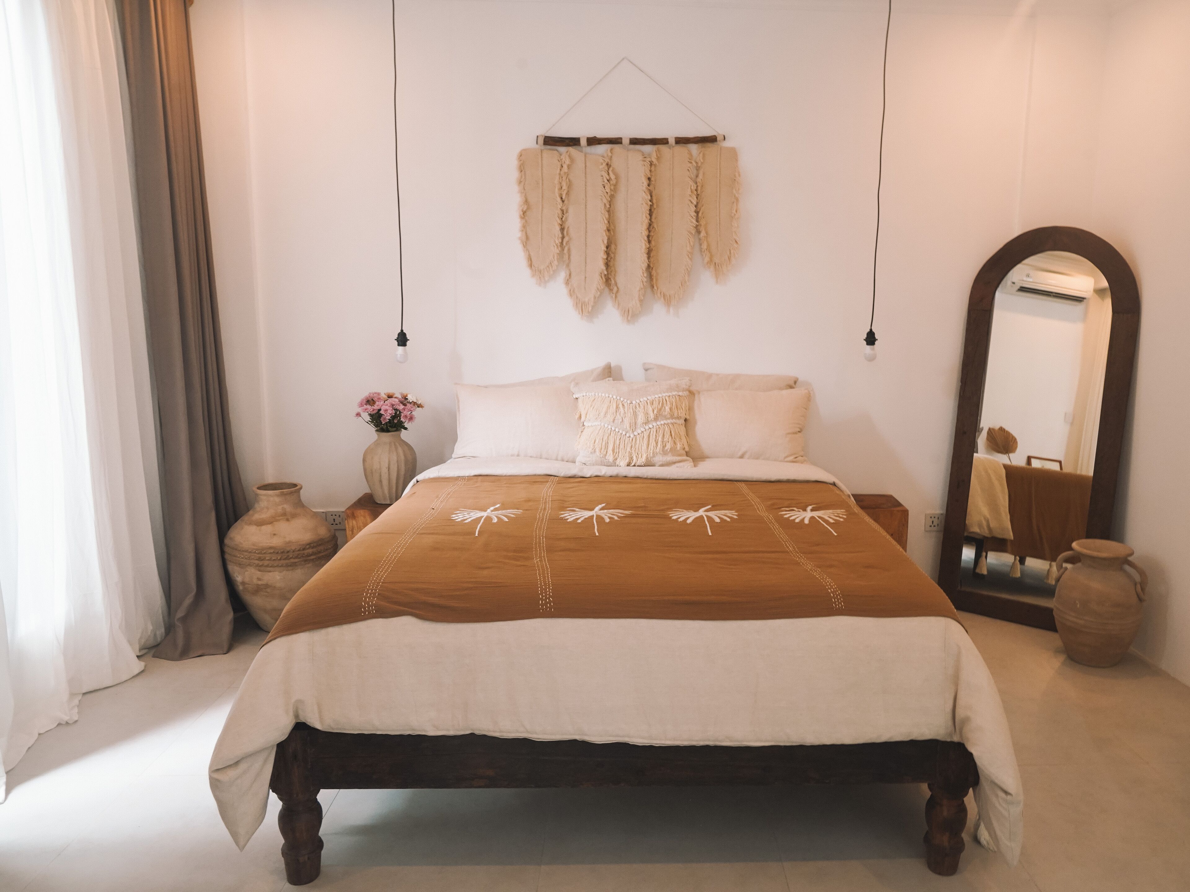A bedroom with a wooden bed, white bedding, a hanging tapestry, a vase with flowers, and a large mirror.