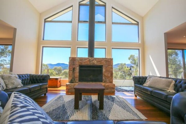 Enjoy a panoramic view of the surrounding landscape from the large windows in this spacious living room, featuring a stone fireplace and comfortable seating areas.