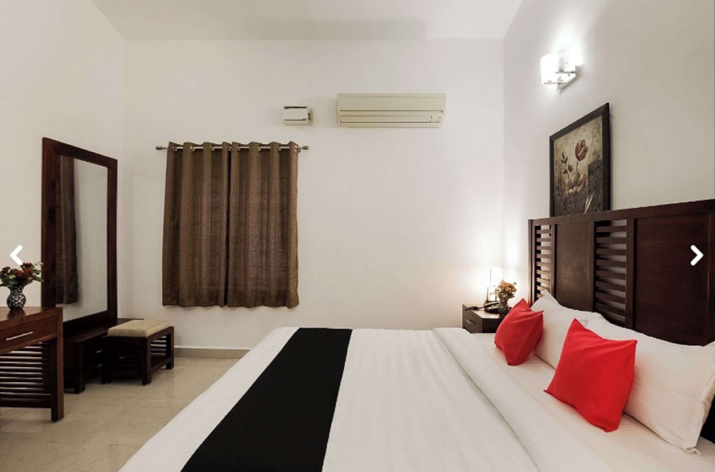 A hotel room with a bed, a desk, a chair, a mirror, a window with curtains, an air conditioning unit, and a wall-mounted light fixture.