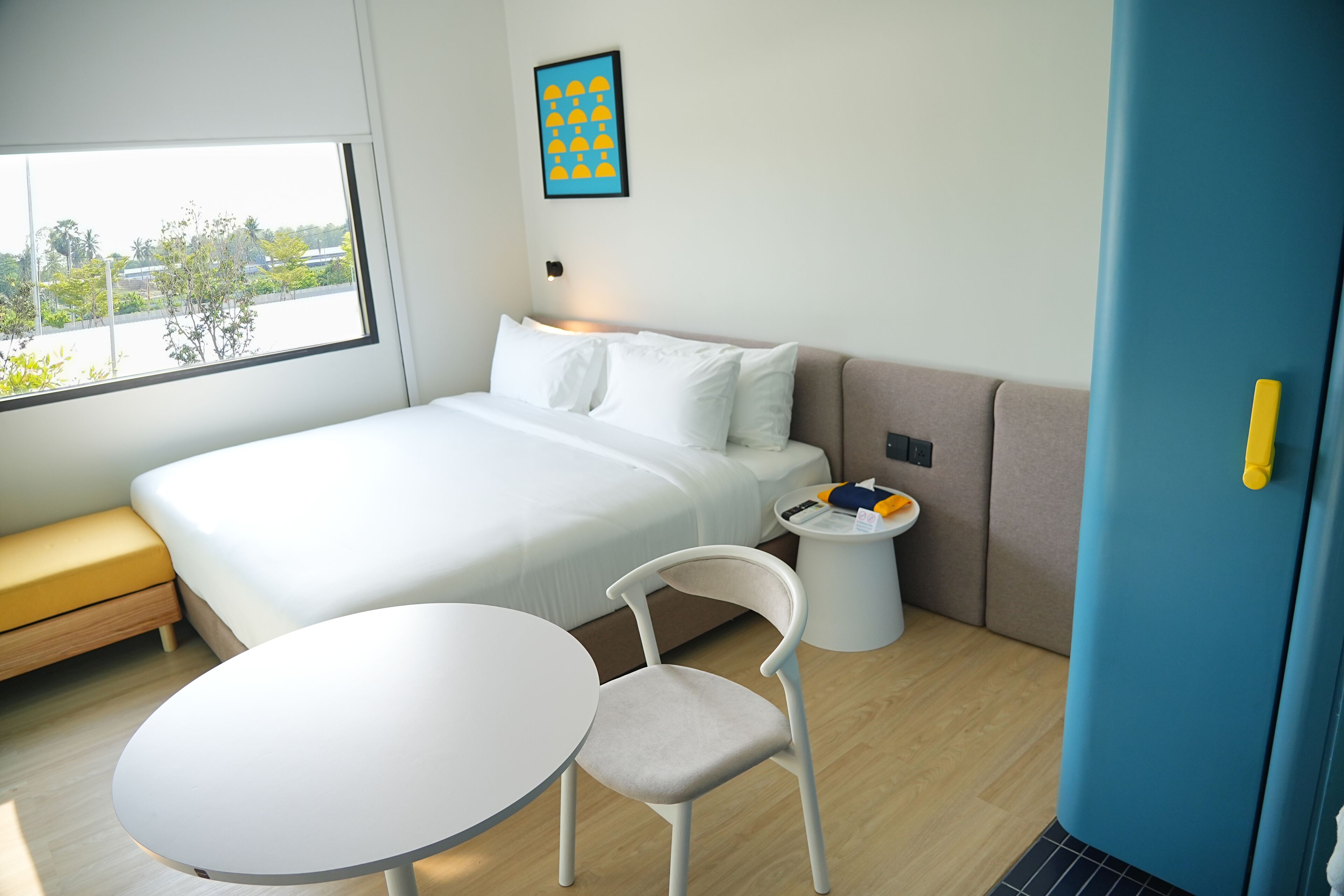 A modern hotel room with a bed, a small round table, a chair, a bedside table with a lamp, and a window with a view of greenery.
