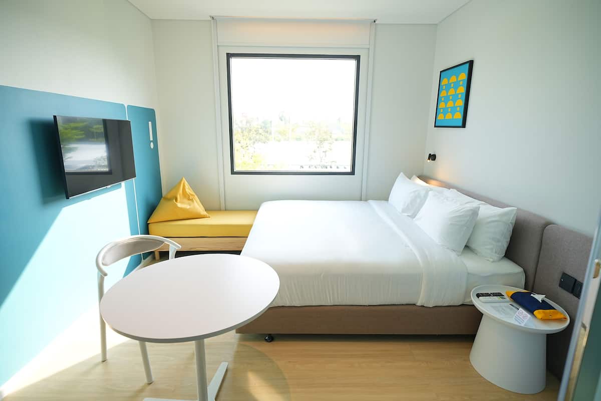 A compact hotel room with a bed, a small table, a chair, a window, and a TV mounted on the wall.