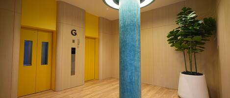 A modern lobby with a wooden floor, a blue pillar, a potted plant, and a yellow elevator.