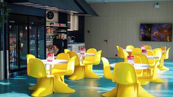Uma área de refeições moderna com cadeiras e mesas amarelas, um balcão de petiscos e uma grande tela de TV na parede.