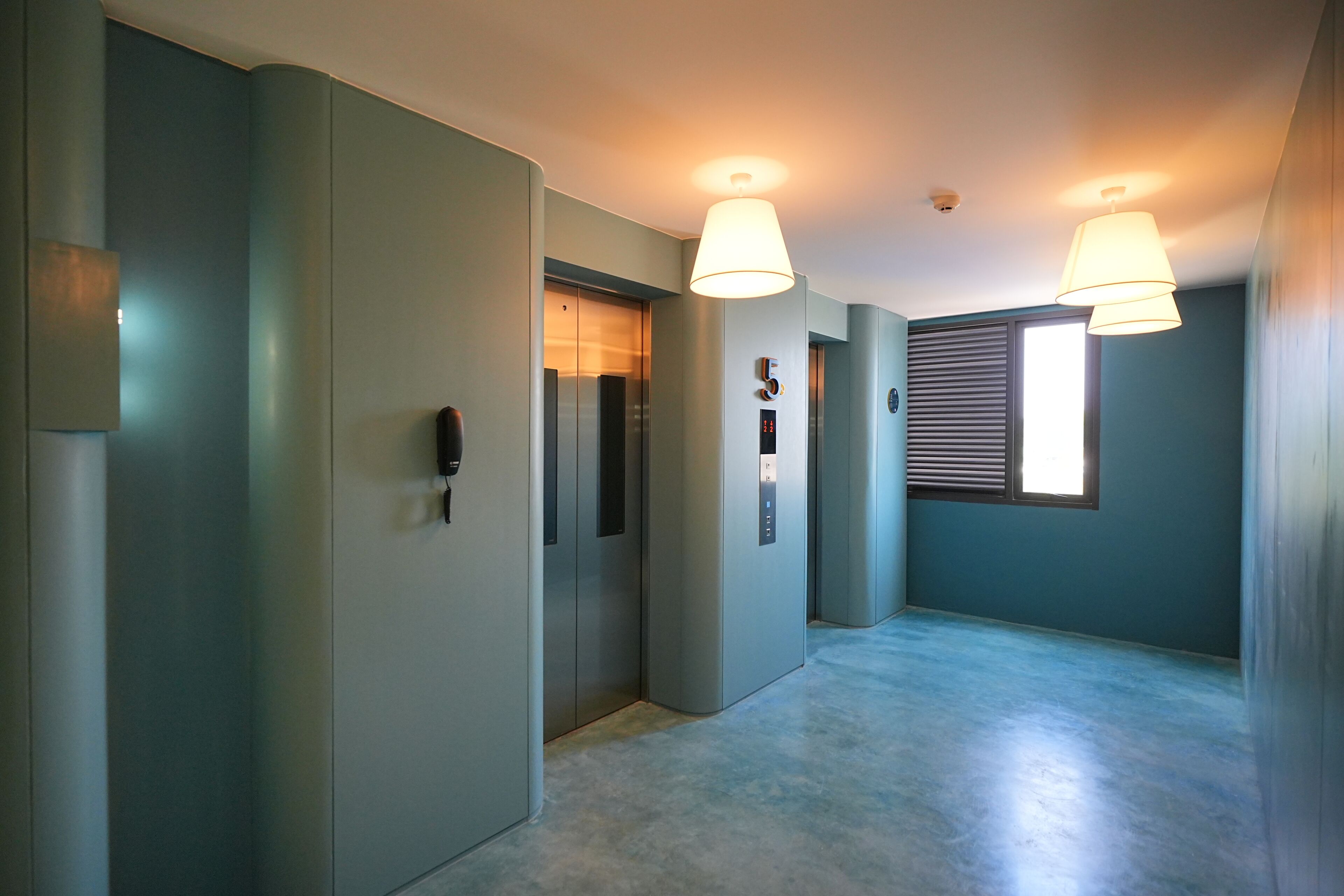 A hallway with two elevators, a window with blinds, and a ceiling light fixture.