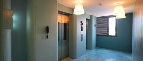 A hallway with two elevators, a window with blinds, and a ceiling light fixture.
