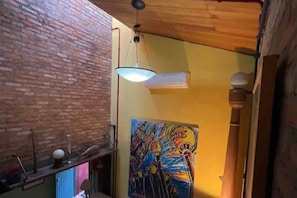 A room with a brick wall, a wooden ceiling, a hanging light fixture, and a colorful painting on the wall. - Ibirapuera Park House (São Paulo)