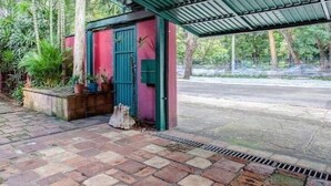 A covered outdoor area with a red and green building, potted plants, and a paved walkway. - Ibirapuera Park House (São Paulo)