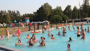 Une piscine où des personnes se baignent et s’amusent, entourée d’arbres et d’une aire de jeux.