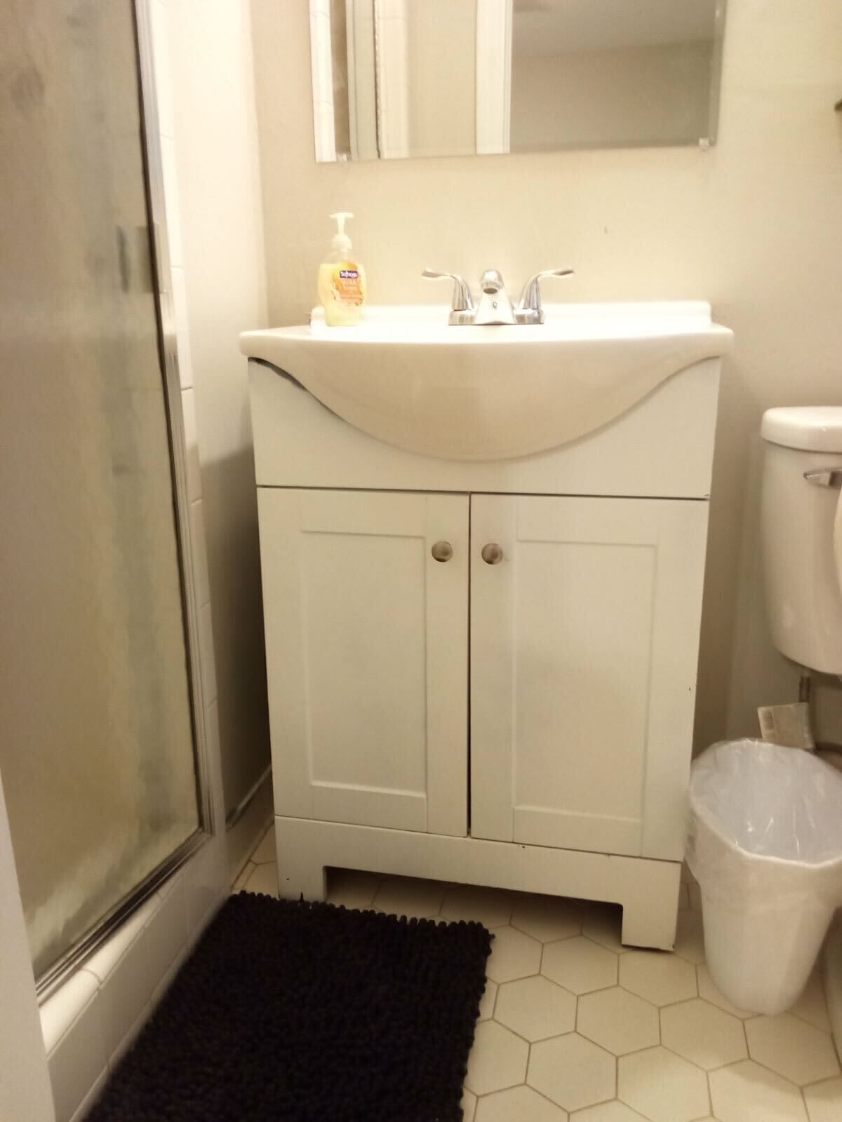 A bathroom with a white sink, mirror, and toilet. There is a black rug on the floor and a trash bin next to the toilet.