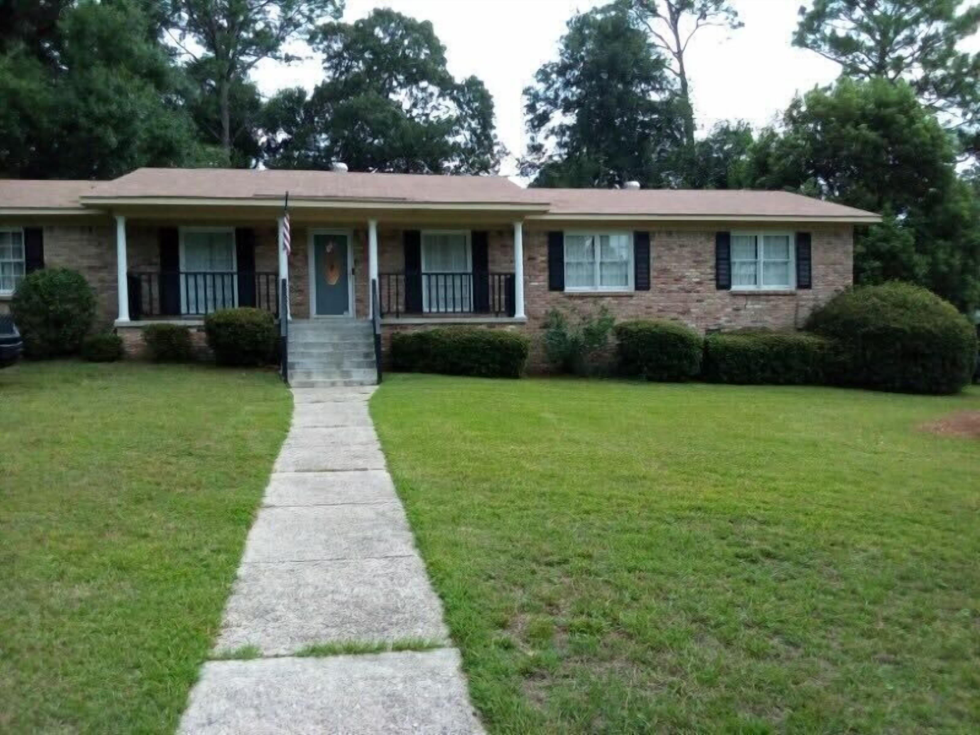 A two-story brick house with a front porch, a front door, and a small front yard with a concrete pathway.