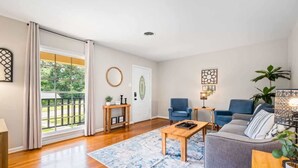 Ein Wohnzimmer mit Holzboden, einem grauen Sofa, einem Couchtisch und einem blauen Teppich mit Muster. Ein Fenster mit Vorhängen, eine Pflanze und Wandbilder sind ebenfalls vorhanden.