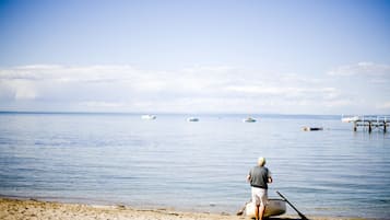 Una platja amb una persona fent caiac, aigua tranquil·la i embarcacions a la distància.