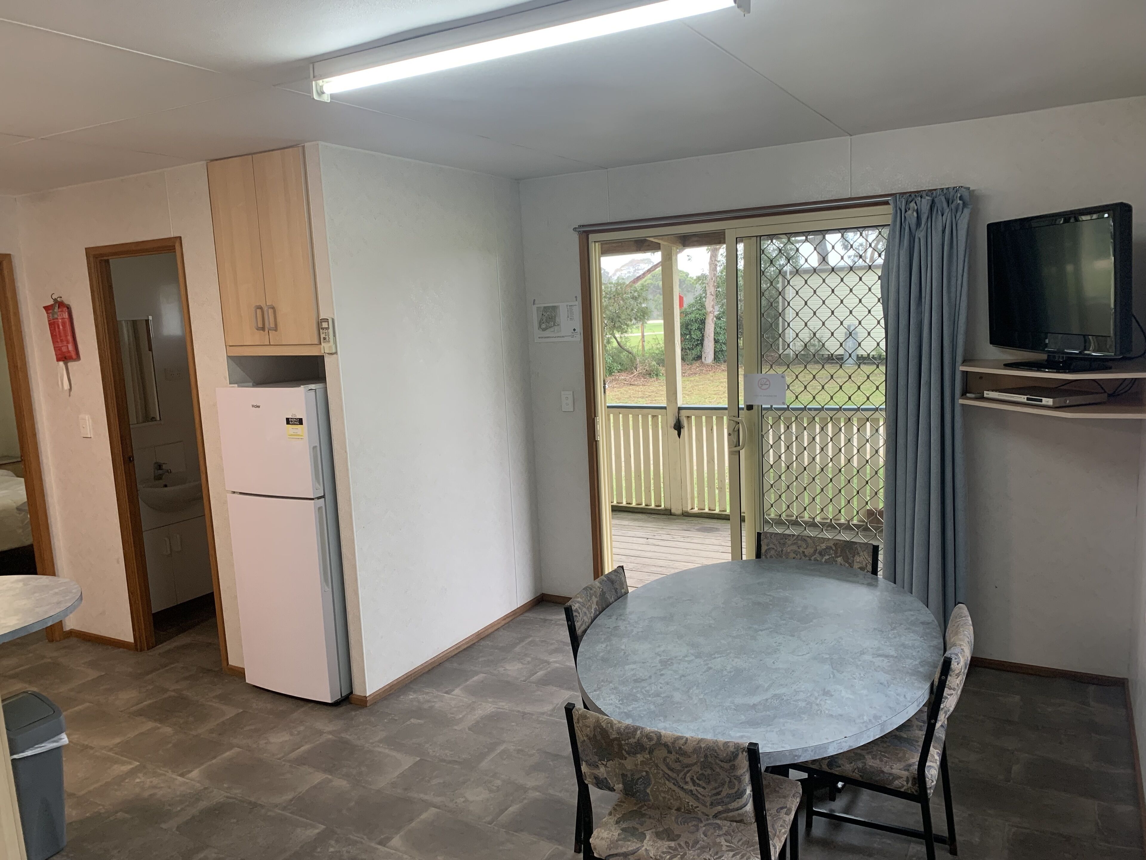 Una cocina compacta con refrigerador, una mesa de comedor con sillas, un televisor en un mueble bajo y una puerta corrediza de vidrio que da a un balcón.