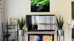 A living room with a TV, two potted plants, a shelf with wine glasses, and a fan. - Cozy house close to all shops/malls, 7 minutes to fort wainwright  (Fairbanks)
