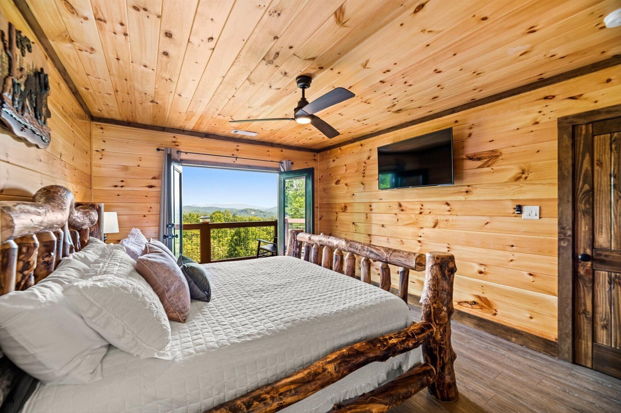 Een slaapkamer in een houten chalet met een bed, een plafondventilator, een aan de muur bevestigde televisie en uitzicht op de natuur.