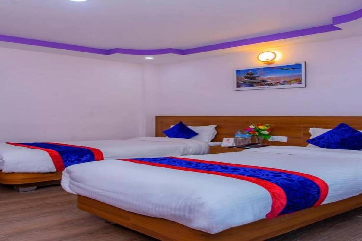 A hotel room with two beds, each with a wooden headboard, white bedding, and blue and red decorative pillows. There is a small table with a vase of flowers and a painting of a pagoda on the wall.