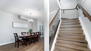 A dining area with a wooden table and chairs, a chandelier, and a potted plant. It also features a staircase with wooden steps and a metal railing. - Central. New Duplex. Sleeps 6. 3 bed/2 bath. (Edmonton)