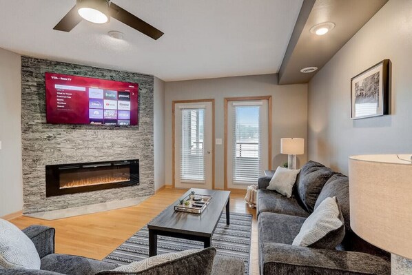 A modern living room with a stone fireplace, a flat-screen TV mounted on the wall, a grey sofa, a coffee table, and a lamp. - WI Dells Lakeside Serenity Condo on Lake Delton - Lake access with boat slip (upon request) (Wisconsin Dells)
